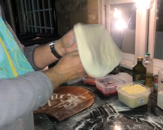 A person's hands tossing and stretching raw pizza dough over a kitchen counter with ingredients.