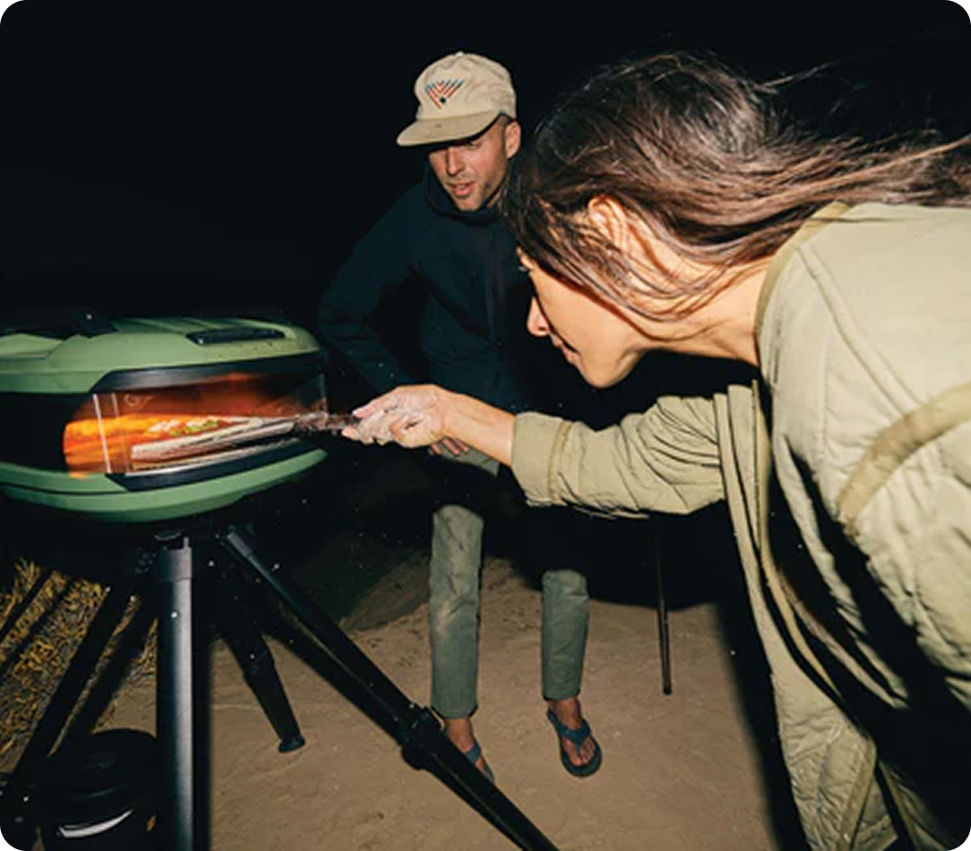 A woman puts a pizza into a portable oven at night as a man looks on.