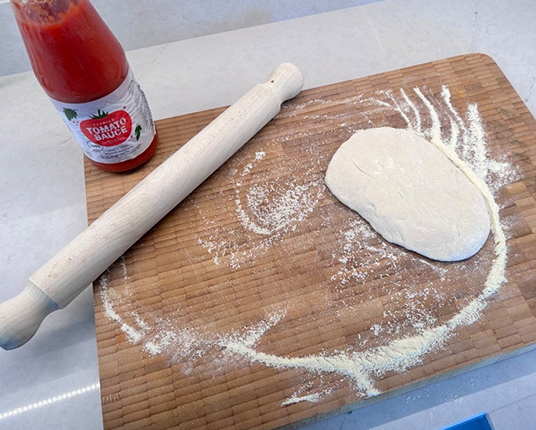 A ball of dough on a floured wooden board with a rolling pin and a bottle of tomato sauce.