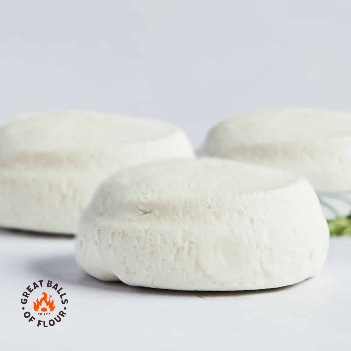 Three balls of raw pizza dough sit on a white surface with a logo in the corner.