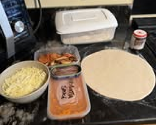 Pizza dough and various toppings in containers laid out on a black countertop, ready for assembly.