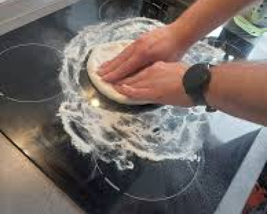 A person cleaning a black glass stove top with a round cleaning pad and white foam.
