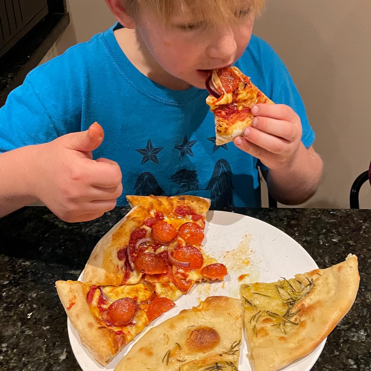 A young boy in a blue shirt eats a slice of pepperoni pizza and gives a thumbs up.
