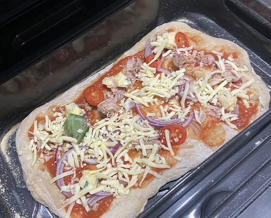 An uncooked rectangular pizza with cheese, tuna, and vegetables on a black baking tray.