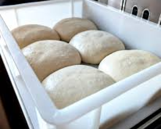 Seven round balls of dough arranged in a white rectangular container, proofing.