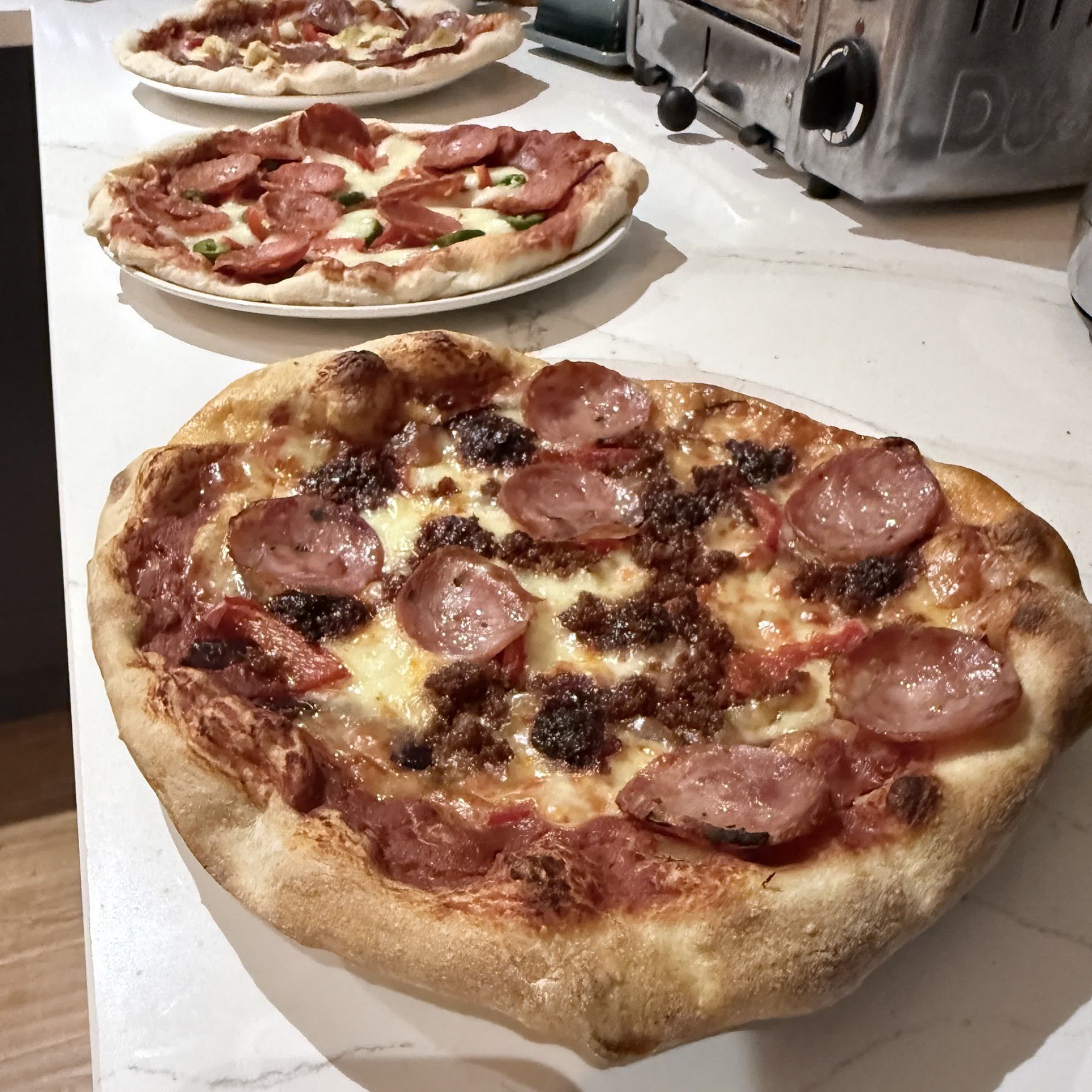 Three freshly baked pizzas with meat toppings sitting in a row on a white marble countertop.