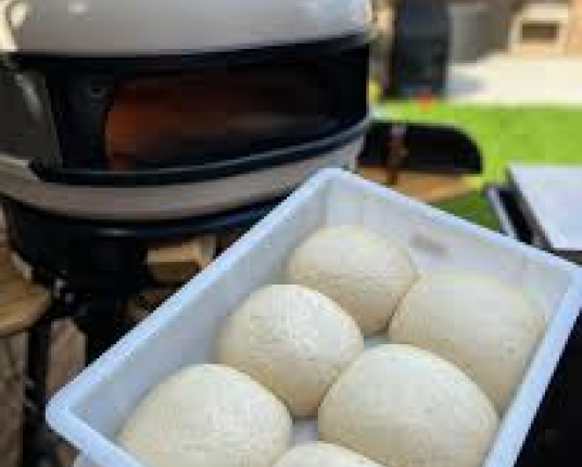 A white tray of pizza dough balls sits in front of an outdoor pizza oven.