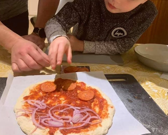 A child and an adult add pepperoni and red onion toppings to a pizza.