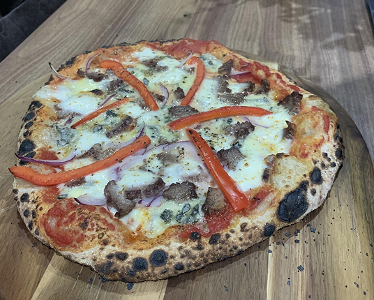 A whole pizza with a charred crust, topped with meat, red peppers, and onions on a wooden board.