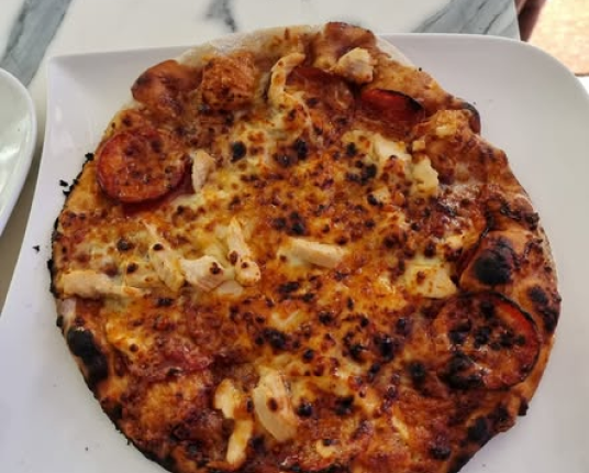 A round pizza with pepperoni, chicken, and a charred crust, served on a square white plate.