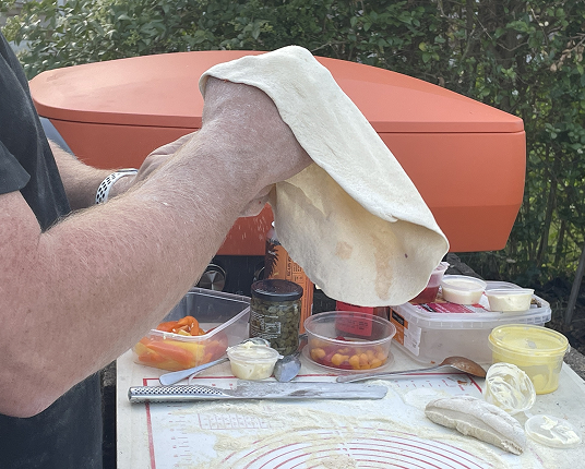 A person stretches pizza dough by hand outdoors, with toppings and an orange pizza oven on the table.
