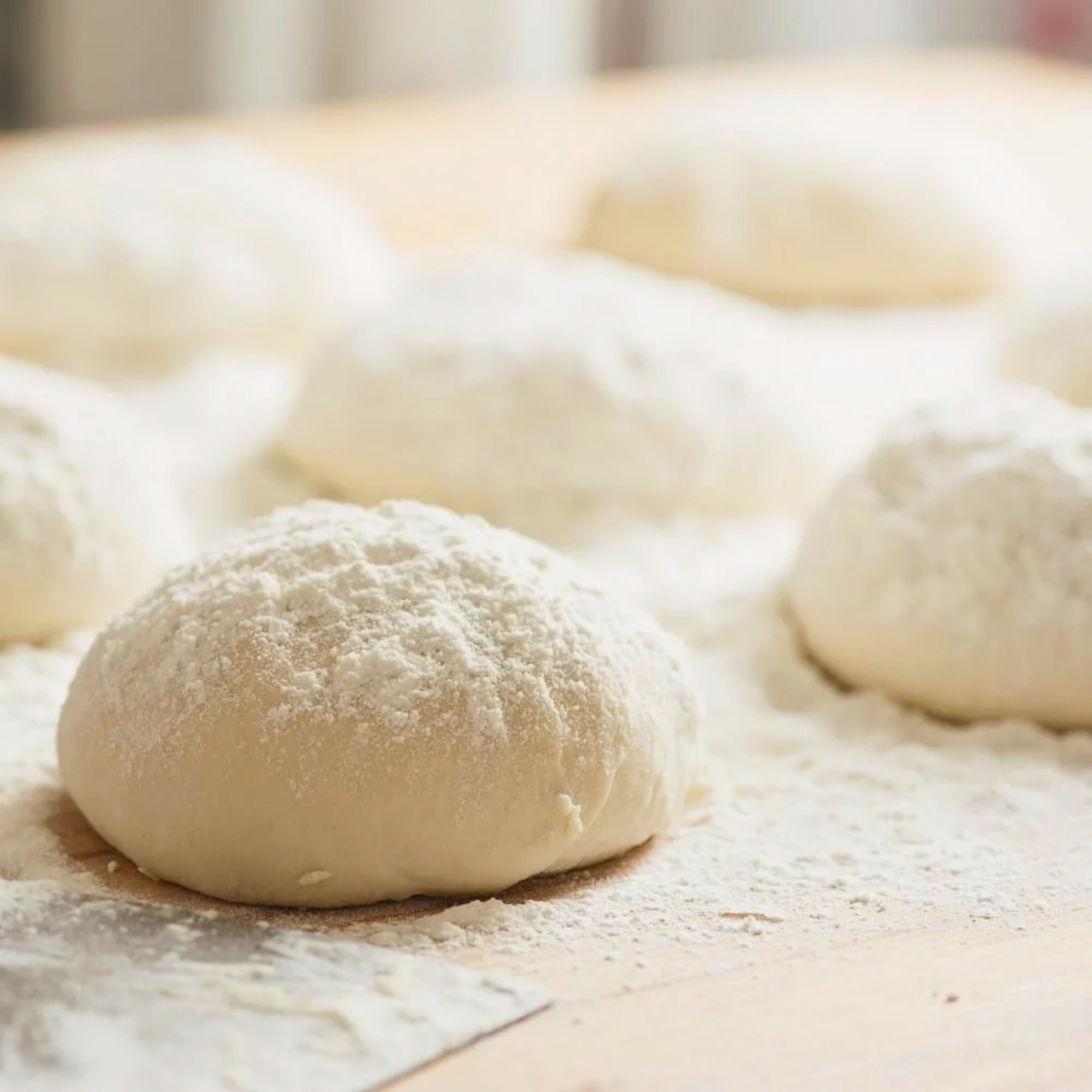 Several balls of raw dough sit on a floured wooden surface, with one in sharp focus.