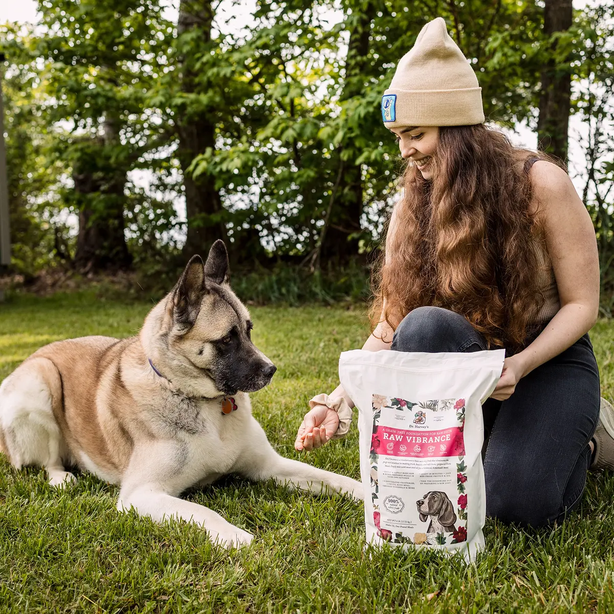 A woman kneels on the grass, holding a bag of dog food and feeding an Akita lying beside her.