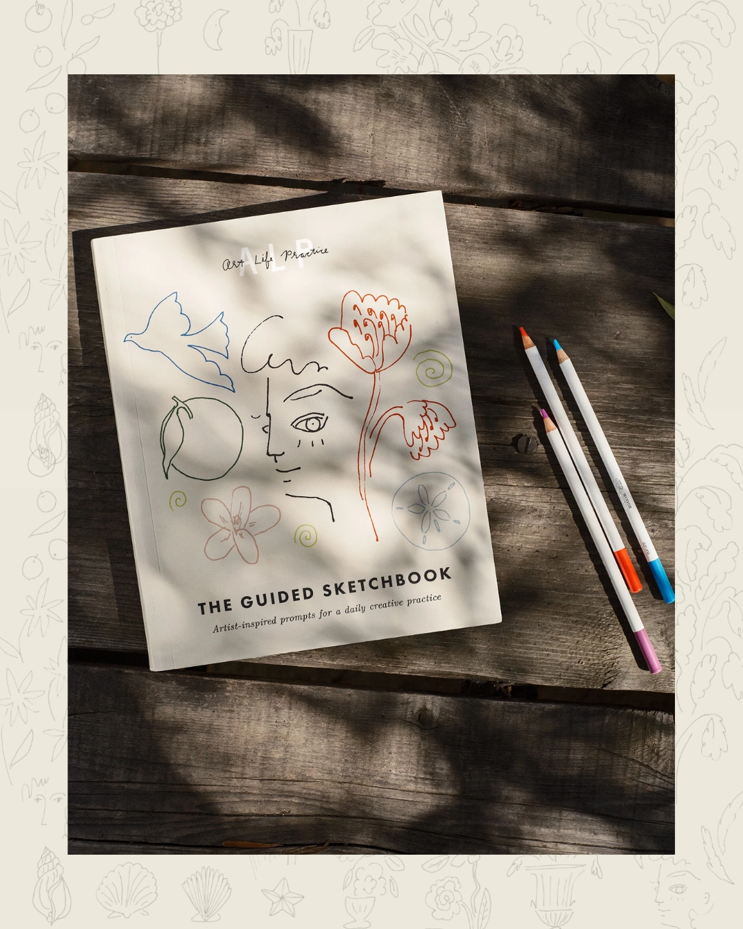 Sketchbook with abstract drawings on a wooden table, three colored pencils beside it.