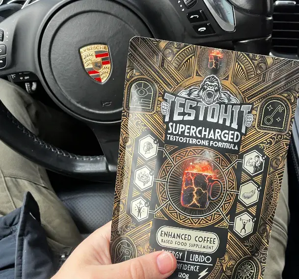 A person in a car holds a packet of TESTOHIT testosterone formula in front of a Porsche steering wheel.