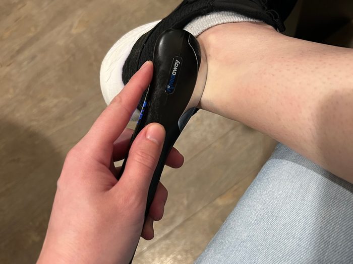 A person holds a black handheld electronic device against their ankle while wearing a shoe and sock.