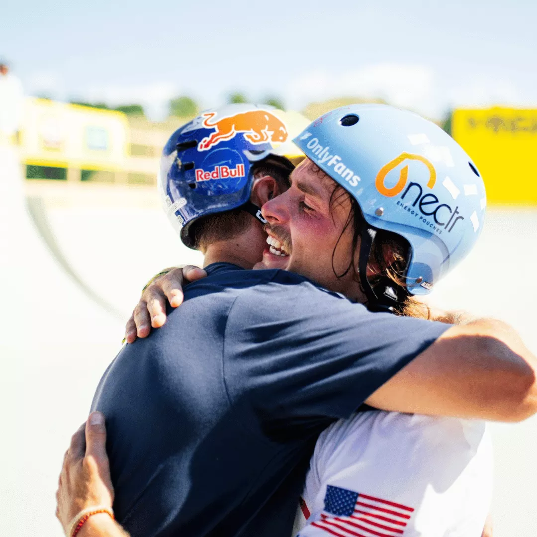 Two people wearing helmets embrace outdoors, logos visible on helmets.