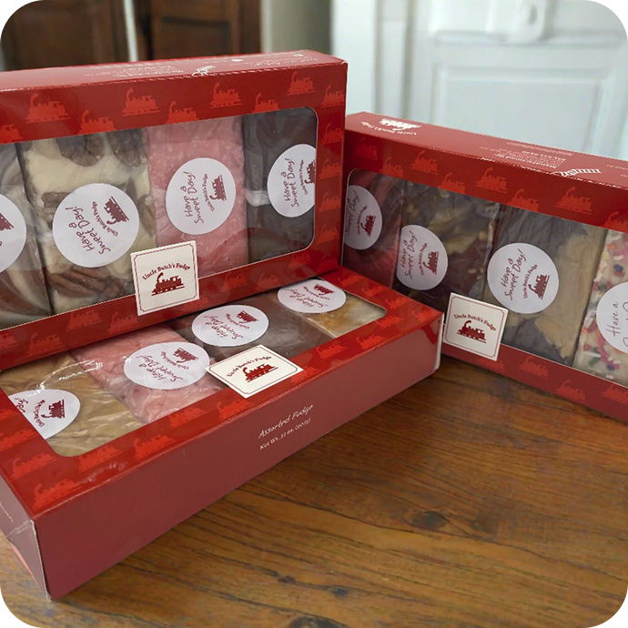 Two red boxes of Uncle Butch's Fudge, filled with assorted flavors, sit on a wooden table.