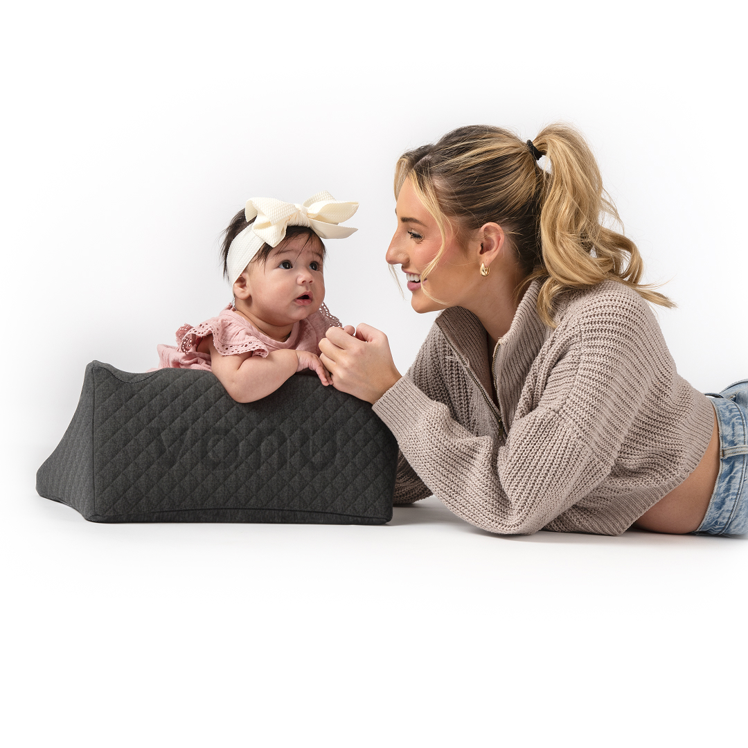 Woman lying next to a baby propped on a cushion, both smiling at each other.