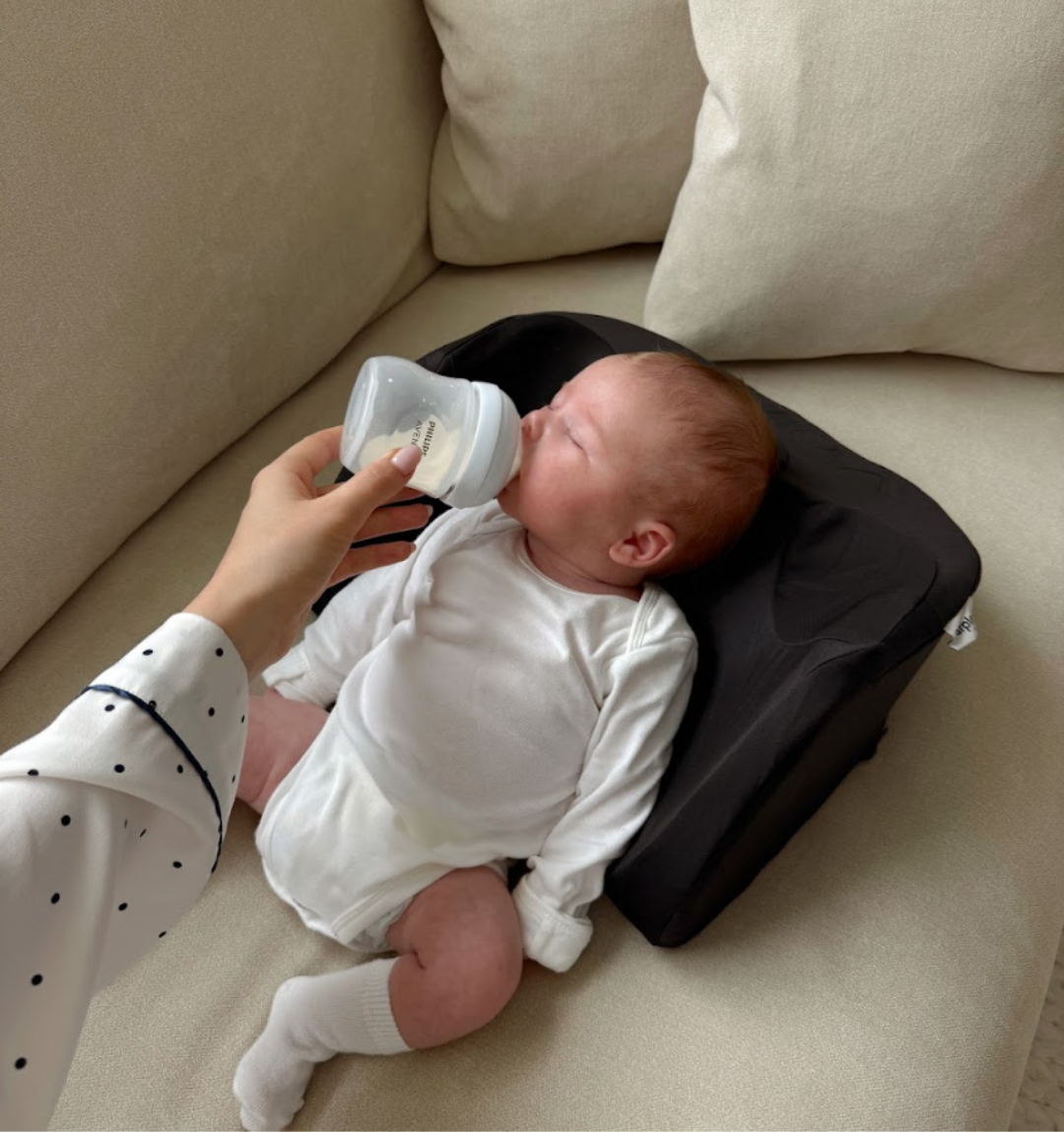 Baby in white outfit being bottle-fed on a beige couch.
