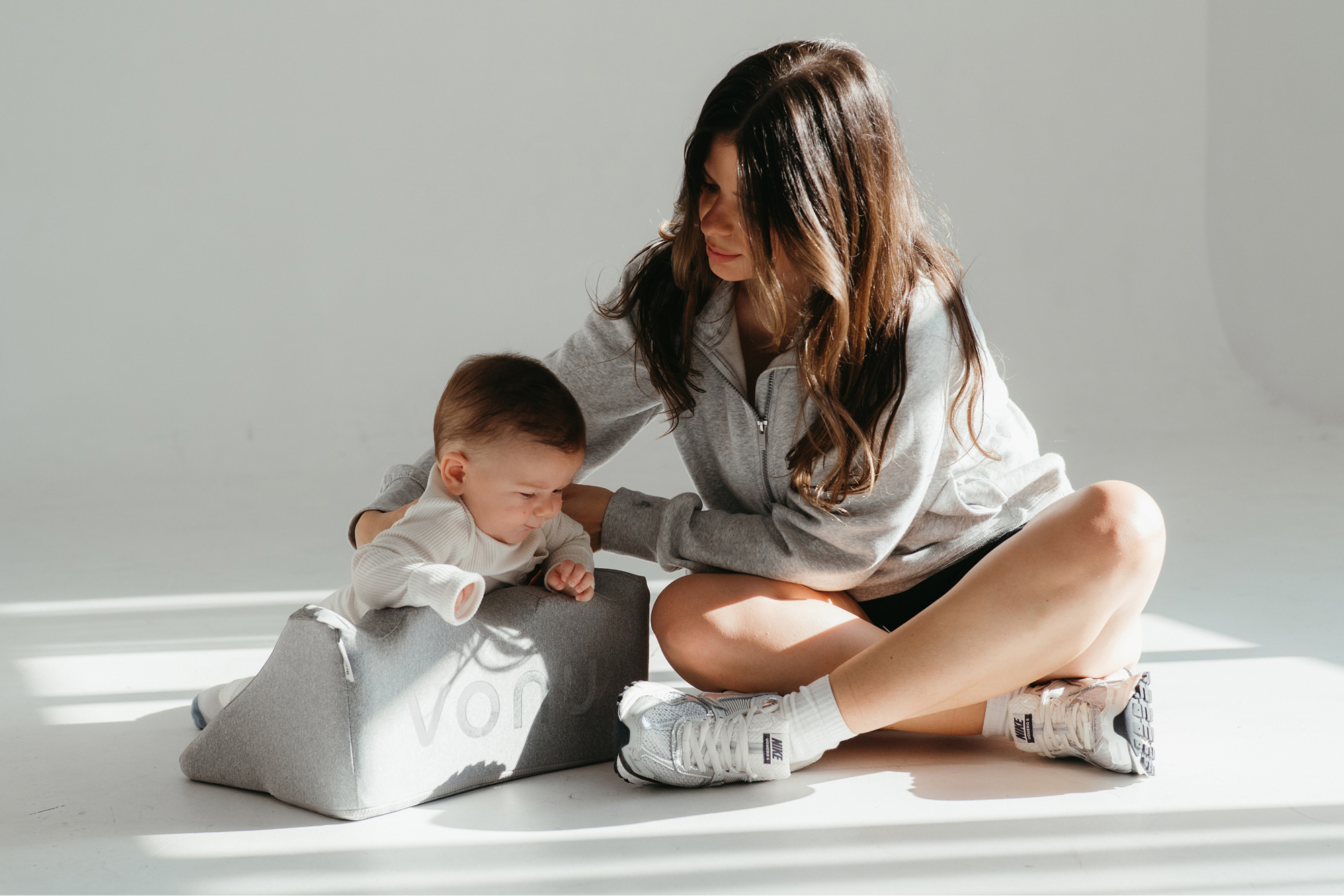 Woman sitting with a baby on a gray cushion in a bright room.