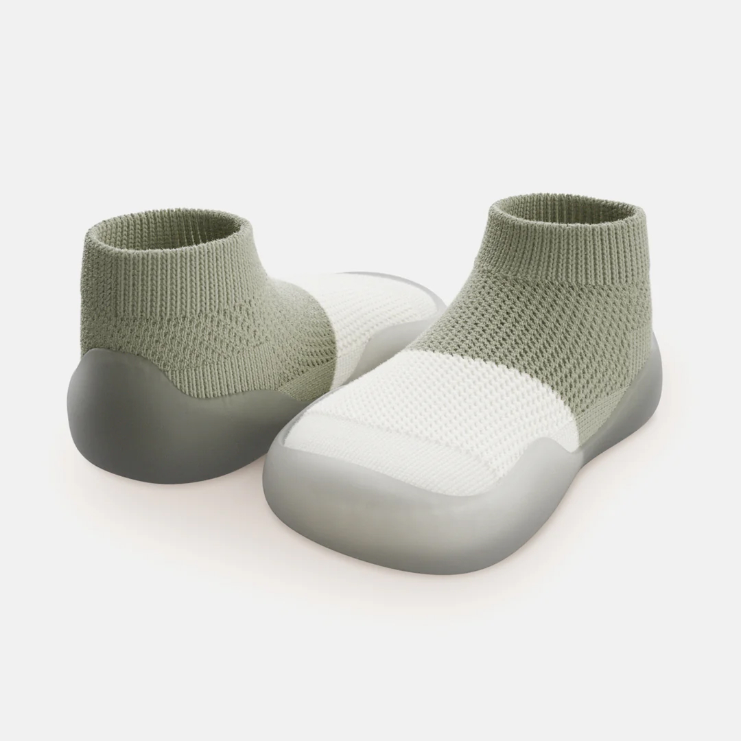 A pair of sage green and white knit sock-shoes with translucent rubber soles on a white background.