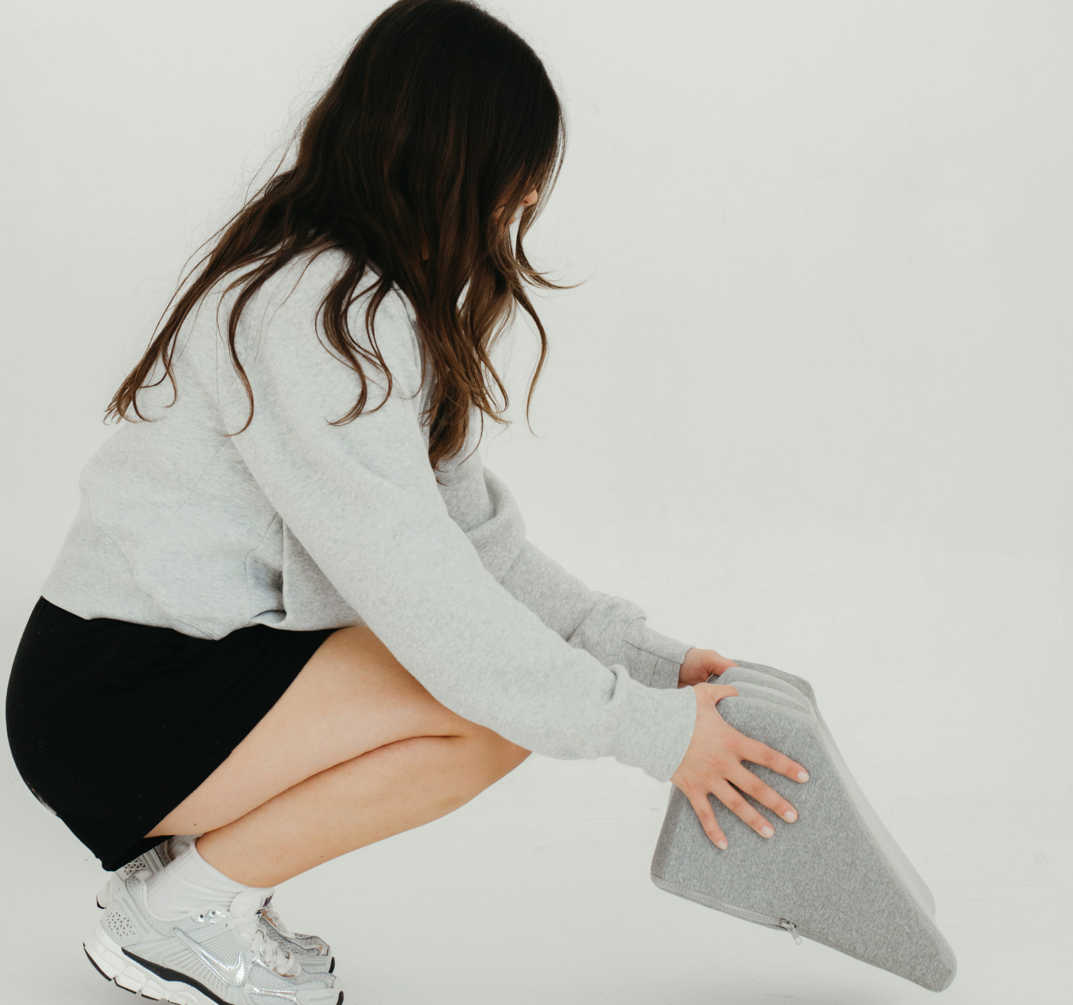 Person wearing a grey sweater squatting, holding a grey triangular cushion.
