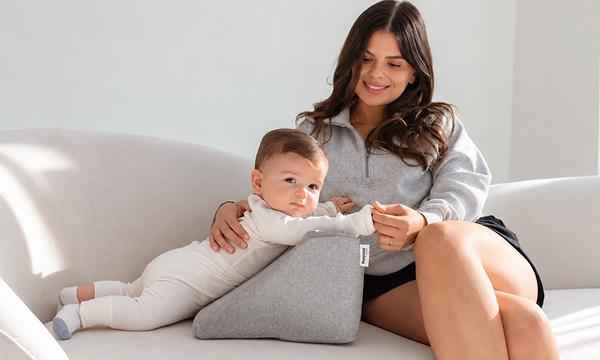 A woman sits on a couch next to a baby lying on its stomach on a grey wedge pillow.