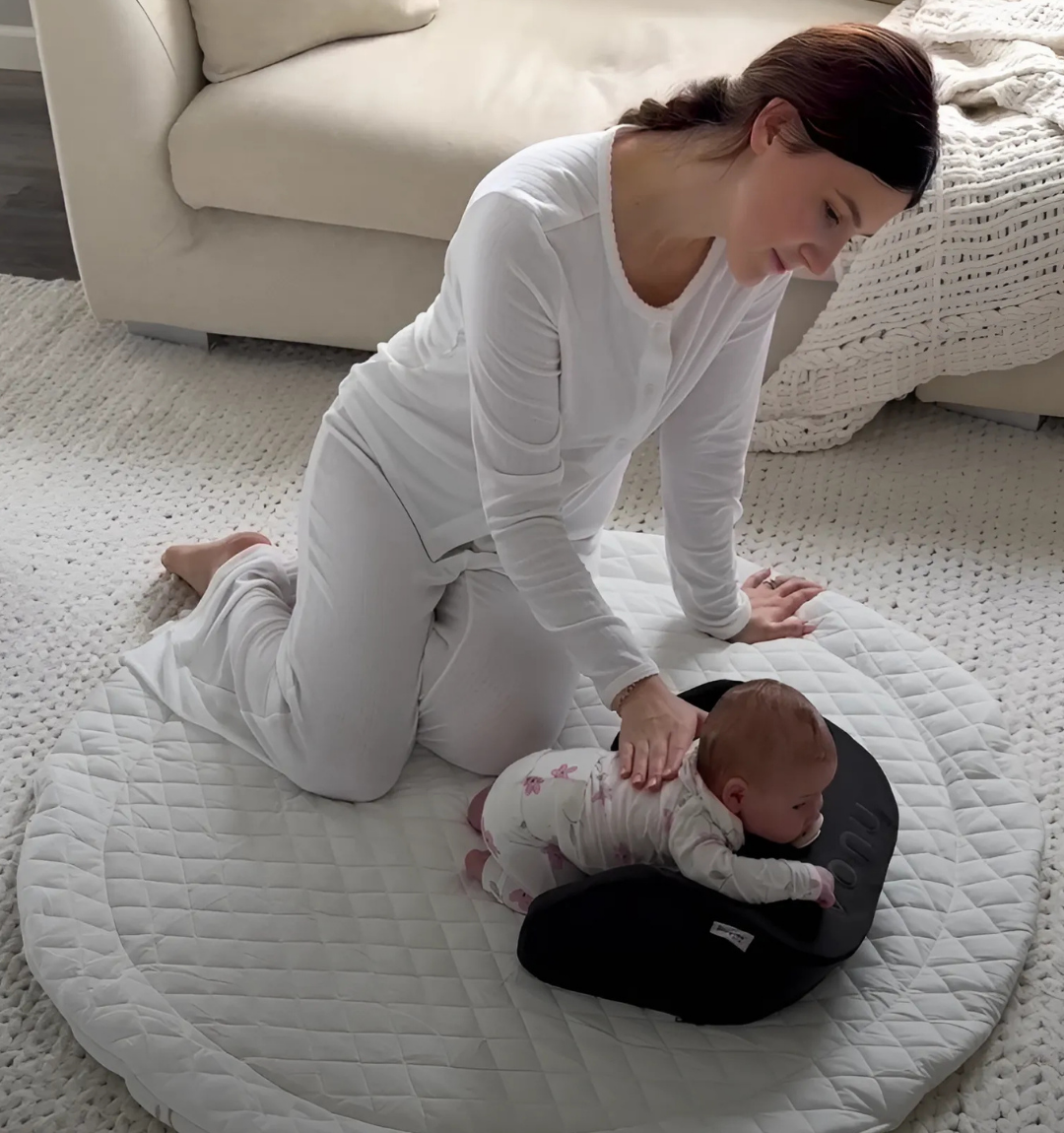 Woman kneeling on a mat, supporting a baby on a cushion.