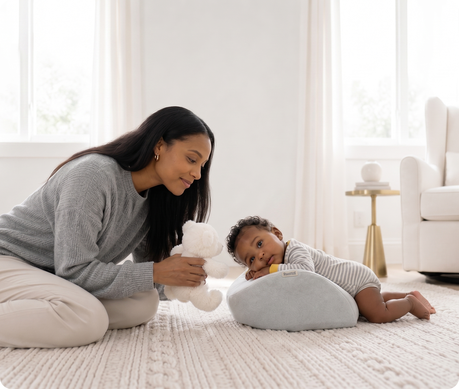 A woman kneels on a rug, holding a teddy bear for a baby doing tummy time on a pillow.