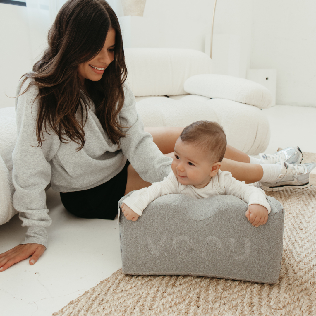Woman sitting with a baby leaning on a gray cushion.