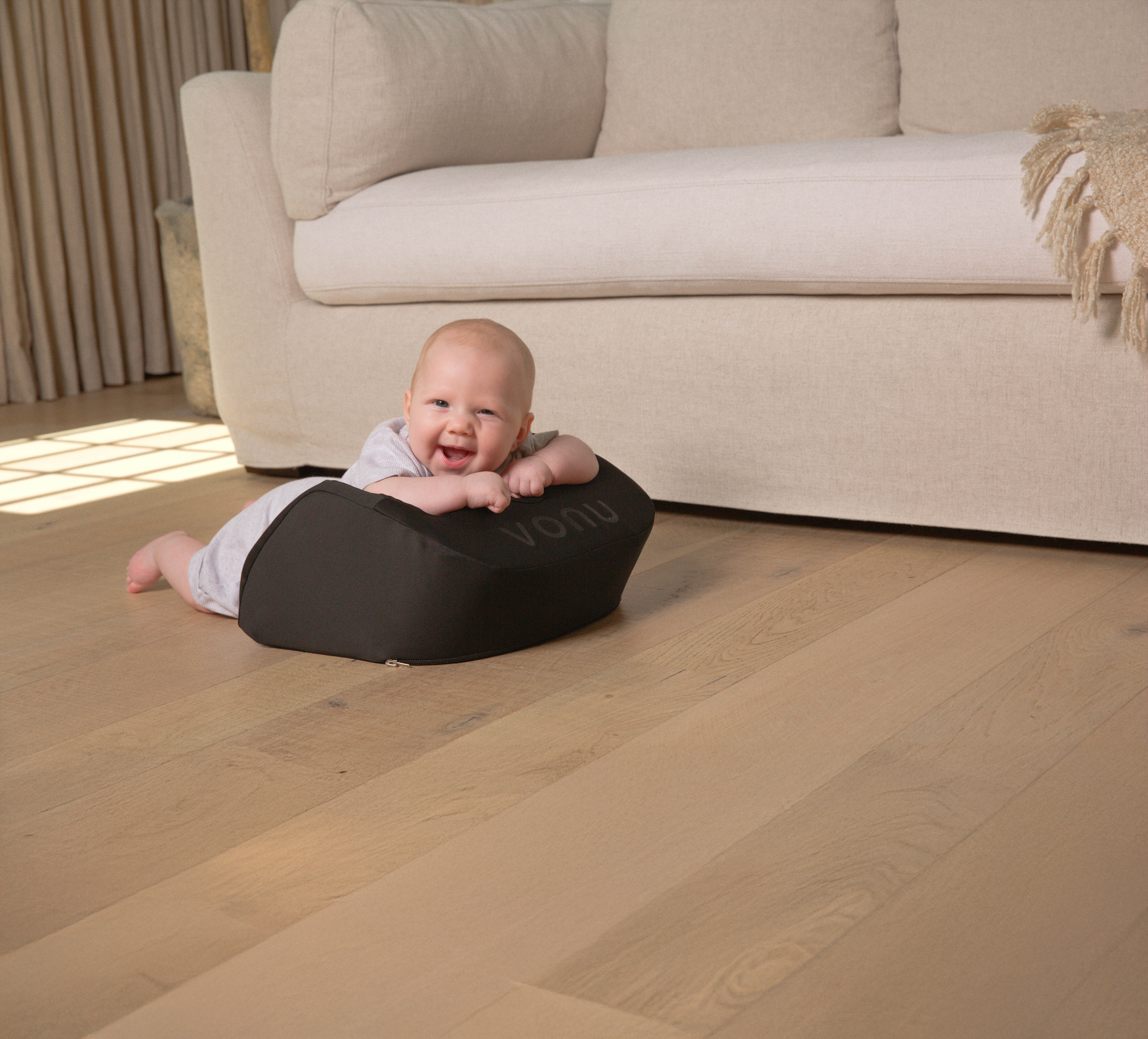 Baby lying on a black cushion on a wooden floor, smiling near a beige sofa.