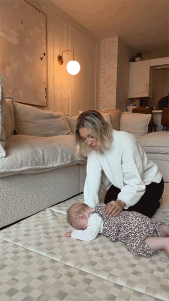 Person assisting a baby lying on a patterned floor mat in a cozy living room.