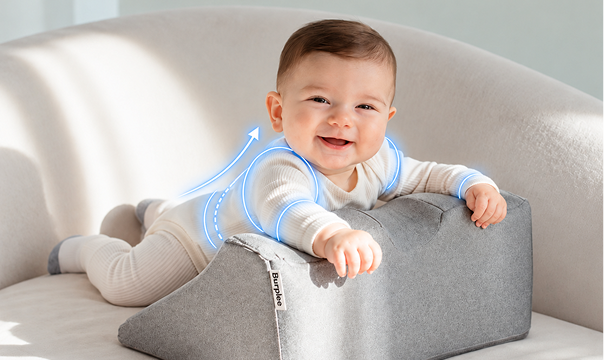 A smiling baby lies on a gray Burplee pillow with a blue graphic indicating back support.