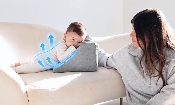A baby lies on a grey wedge pillow on a couch as a woman looks on.