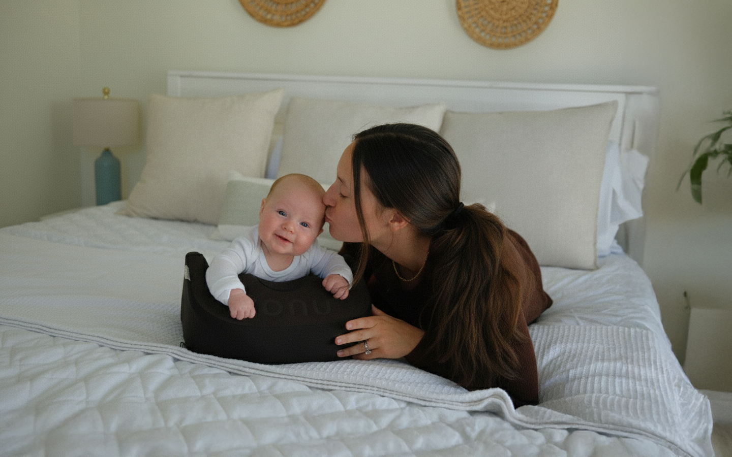 A woman lies on a bed, kissing a smiling baby who is propped up in a lounger.