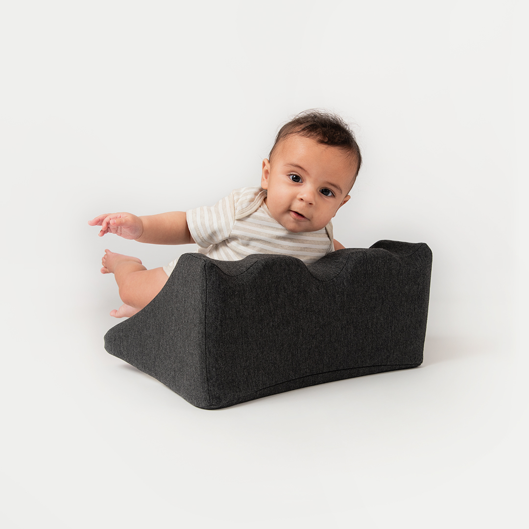 Baby seated in a gray support pillow against a white background.