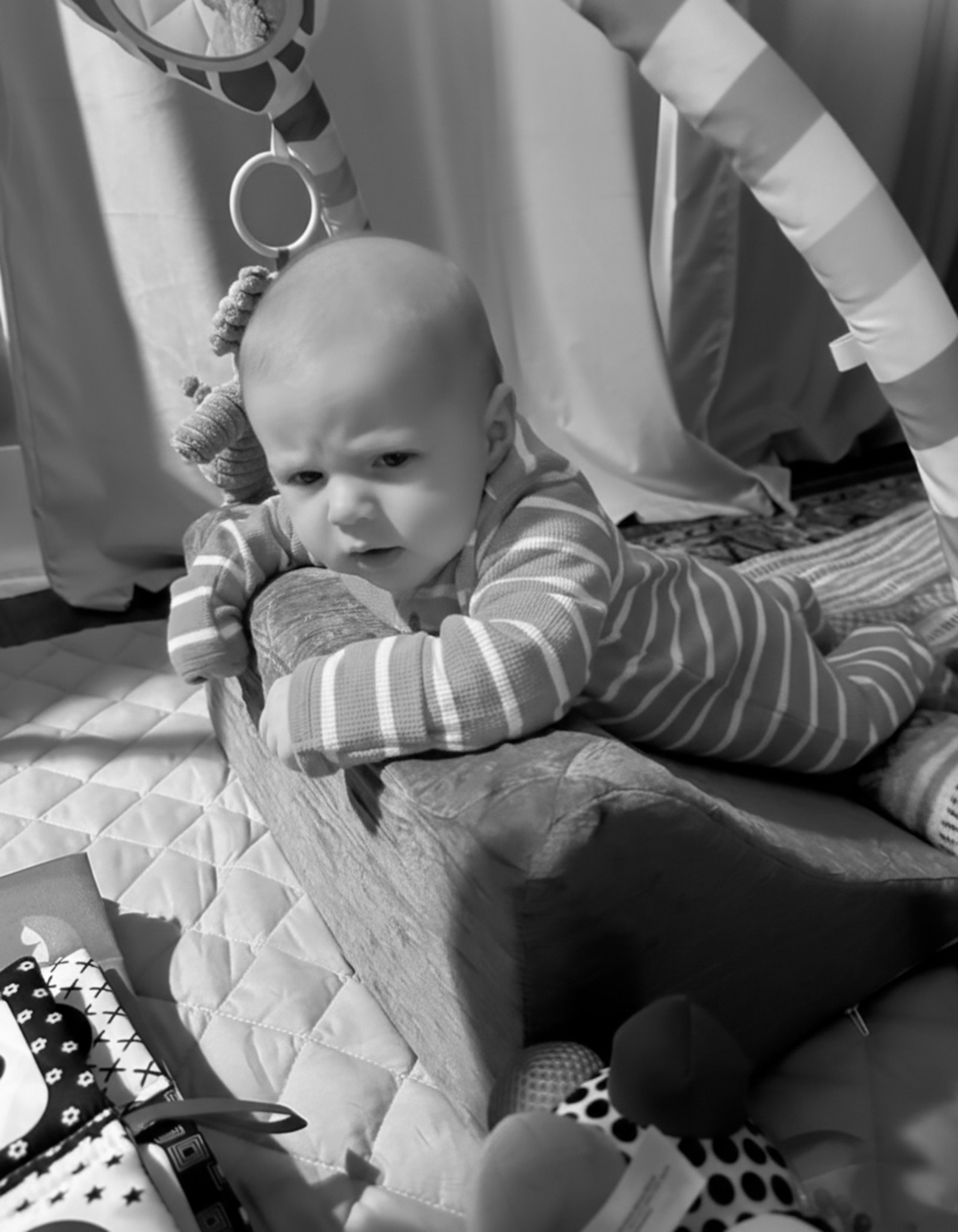 A black and white photo of a baby lying on its stomach on a quilted playmat.