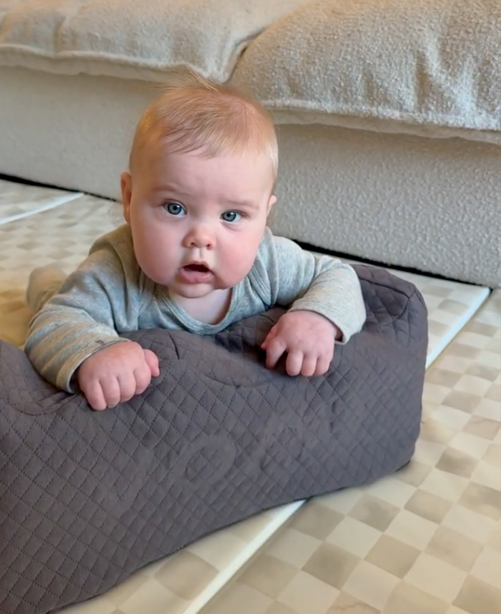 Baby lying on a soft, quilted, gray cushion indoors.