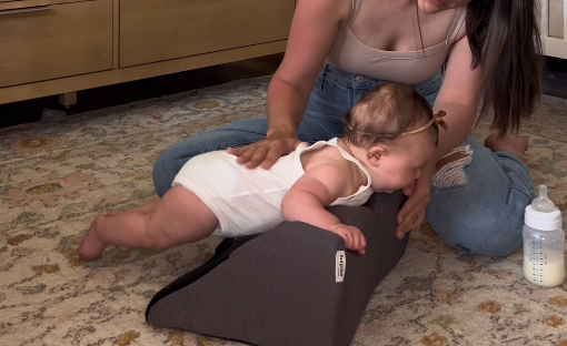 A woman assists a baby doing tummy time on a grey wedge pillow on the floor.