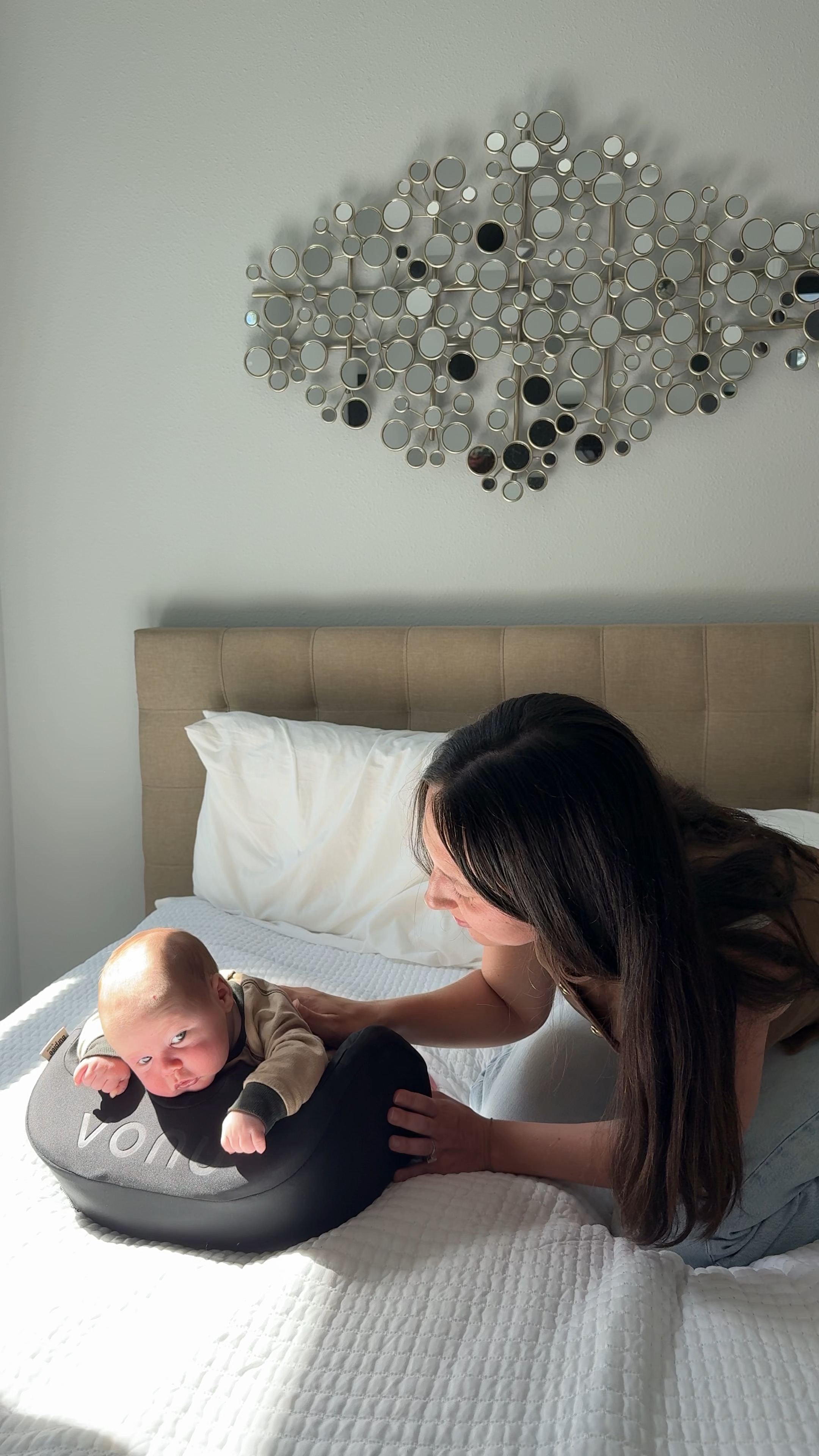 Baby lying on a cushion with a woman leaning over in a well-lit bedroom.