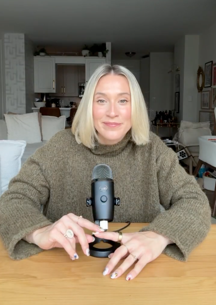Person sitting at a table with a microphone, wearing a brown sweater.