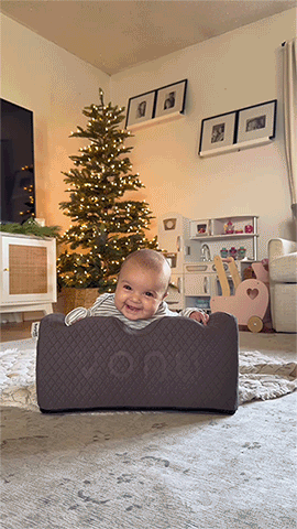 Smiling baby supported by cushion in cozy room with Christmas tree.