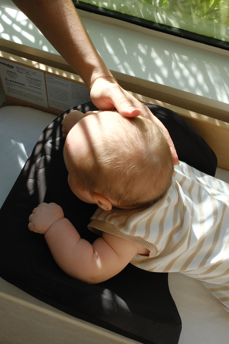 Infant lying on a mat with an adult's hand resting on its head.