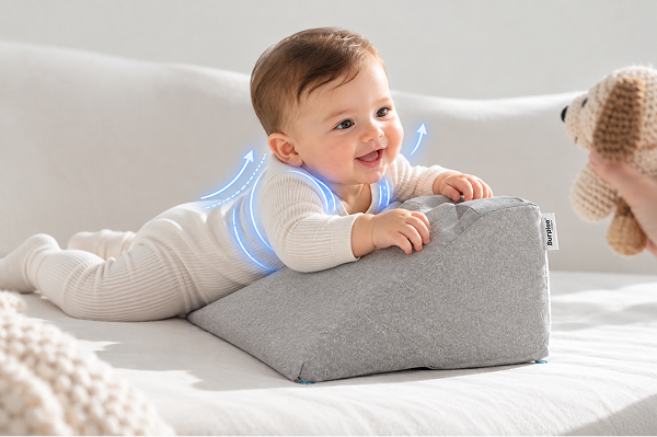 A smiling baby lies on a grey wedge pillow, looking at a person holding a stuffed dog toy.
