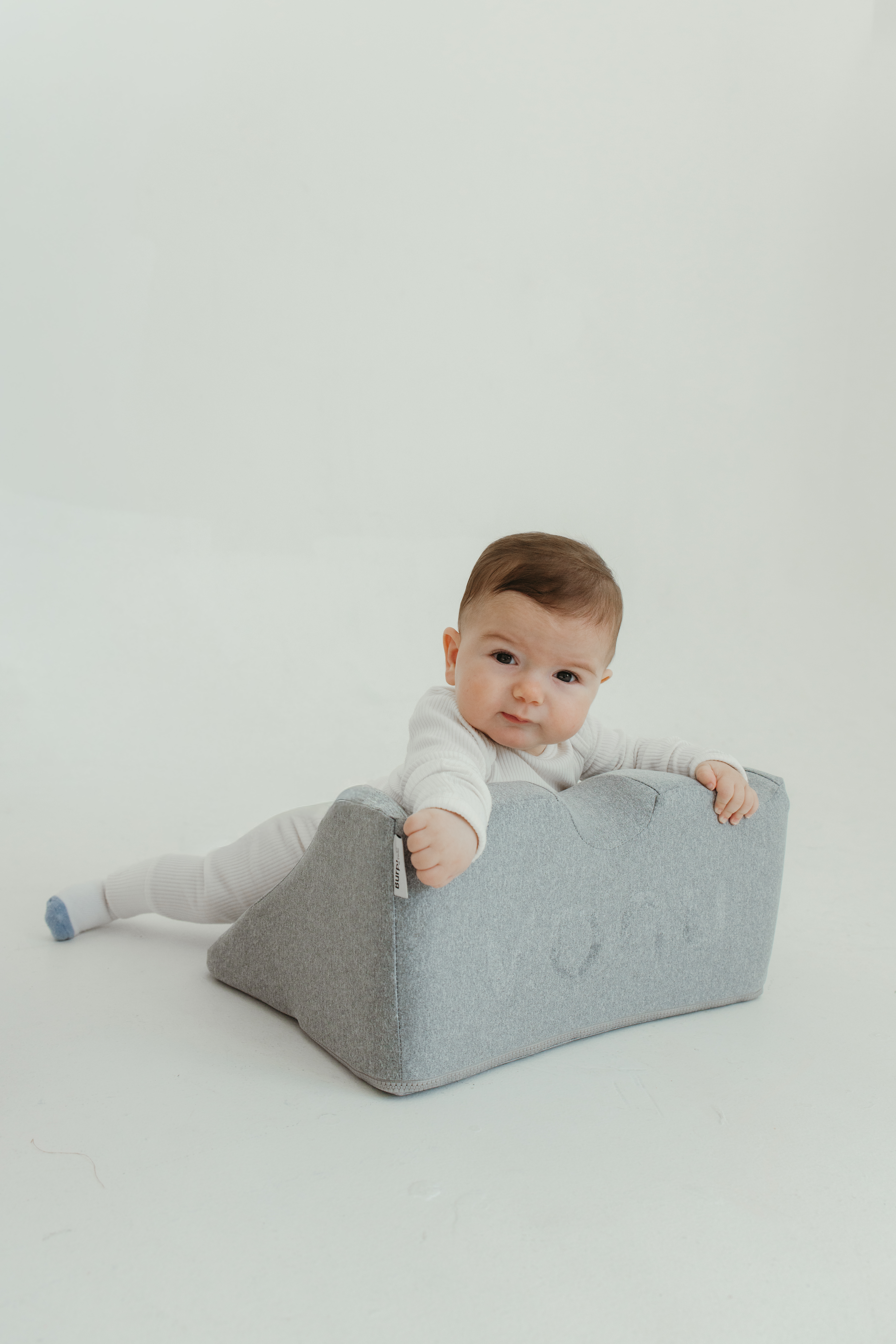 Baby in white outfit lying on a gray cushion on a white background.