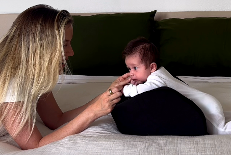 A woman lies on a bed, looking at a baby who is propped up on a black pillow.