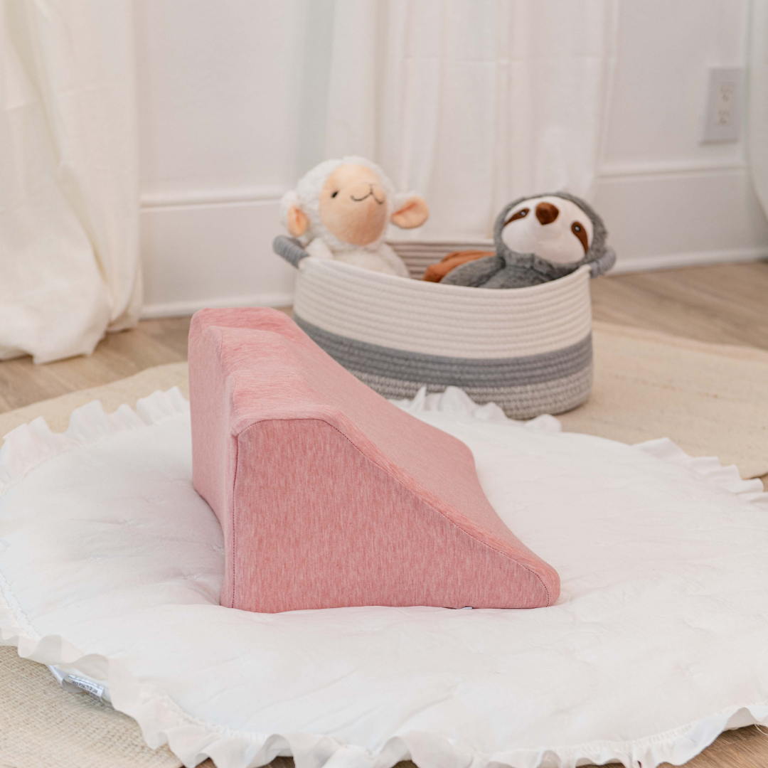Pink wedge pillow on white mat with stuffed animals in a basket.