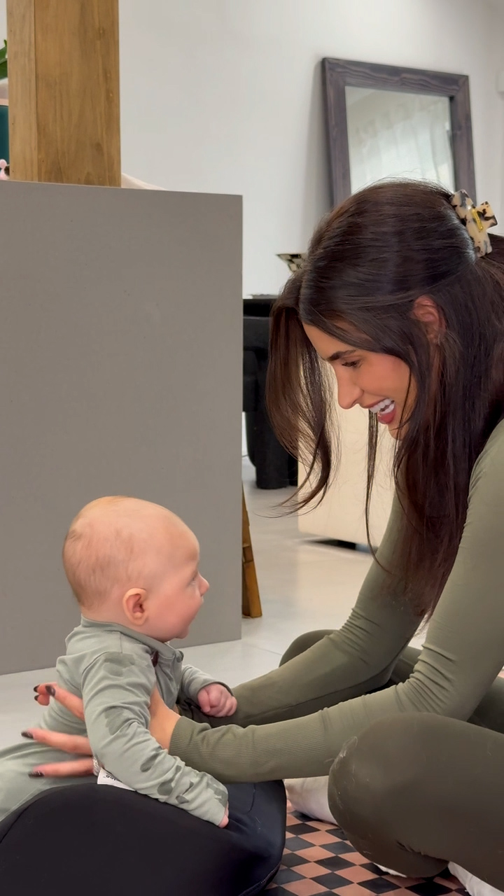 Woman smiling at a seated baby indoors.