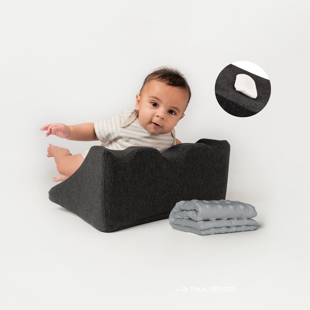Baby in striped outfit sitting in a dark foam support with gray blankets nearby.
